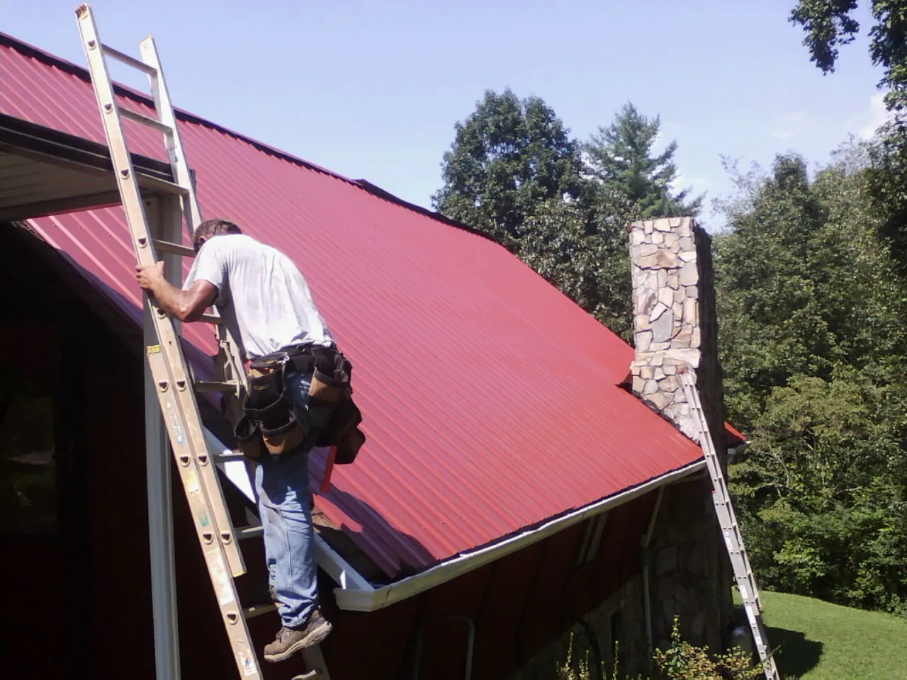 Roofer on a ladder beside a red metal roof and stone chimney — Soffit & Fascia Repair by Roofing Brothers in North Smithfield